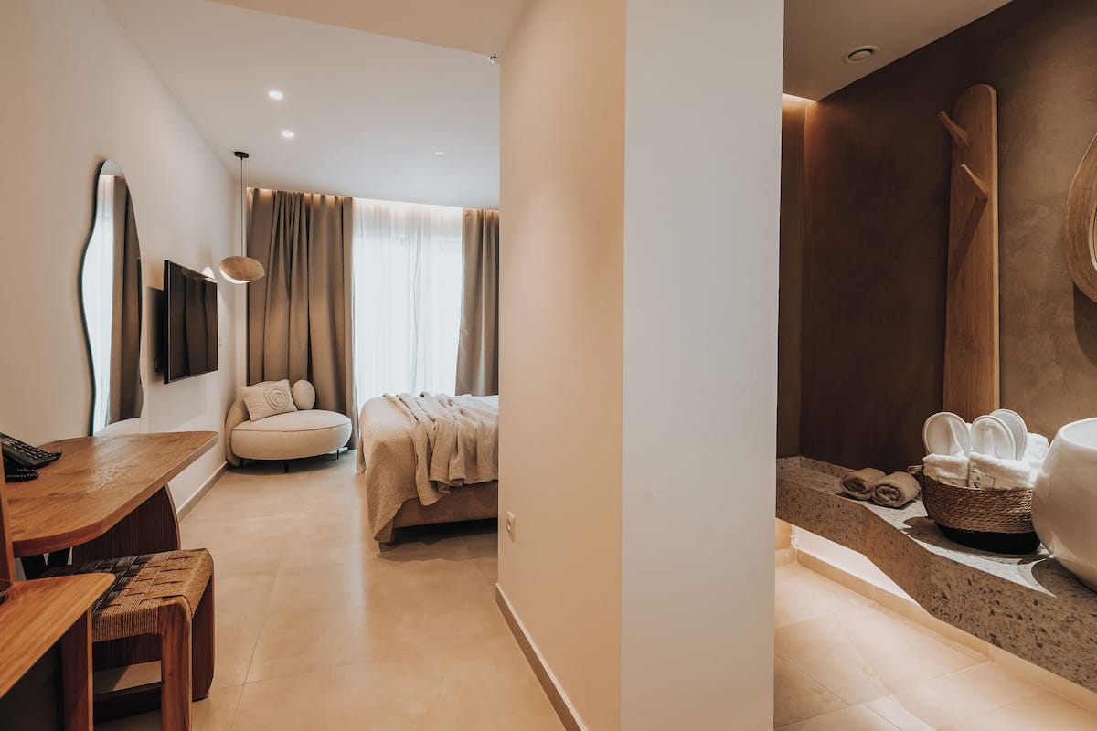 A modern hotel room with a wooden desk, a bed with a beige blanket, a large mirror, and a bathroom with a wooden bath mat and towels.