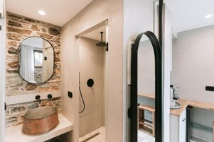 Baño con pared de piedra, espejo redondo, lavamanos de cobre, zona de ducha y encimera de madera.