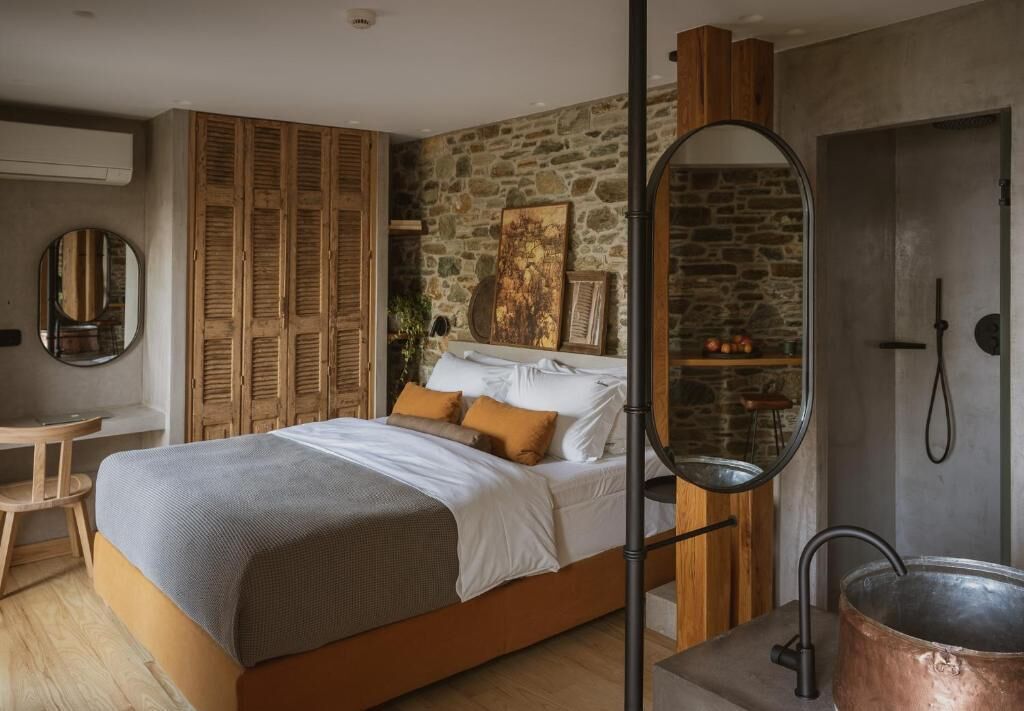 Une chambre avec un mur en pierre, une armoire en bois, un lit avec des oreillers, un miroir rond et une salle de bain avec une douche et un pot en cuivre.