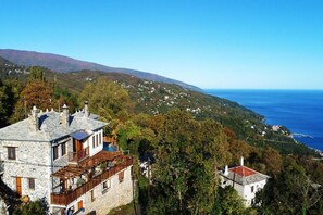A stone building with a balcony, surrounded by trees and overlooking a coastal area. - Adama (Zagora-Mouresi)