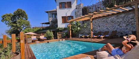 Una piscina con terraza de madera y sillones de descanso, pared de piedra y un edificio de dos plantas con balcón.