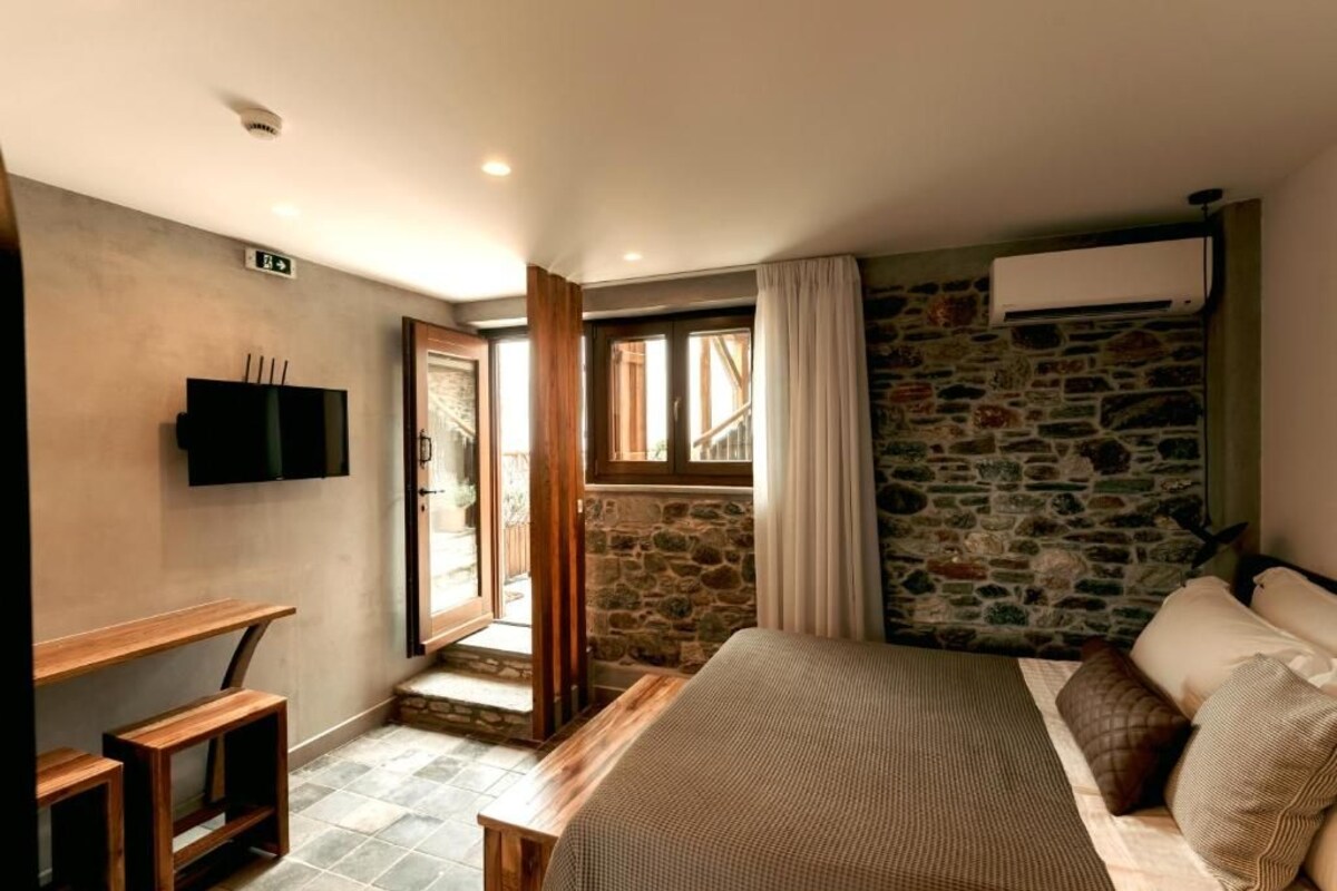 A bedroom with a stone wall, a bed with pillows, a TV on the wall, and a wooden bench.
