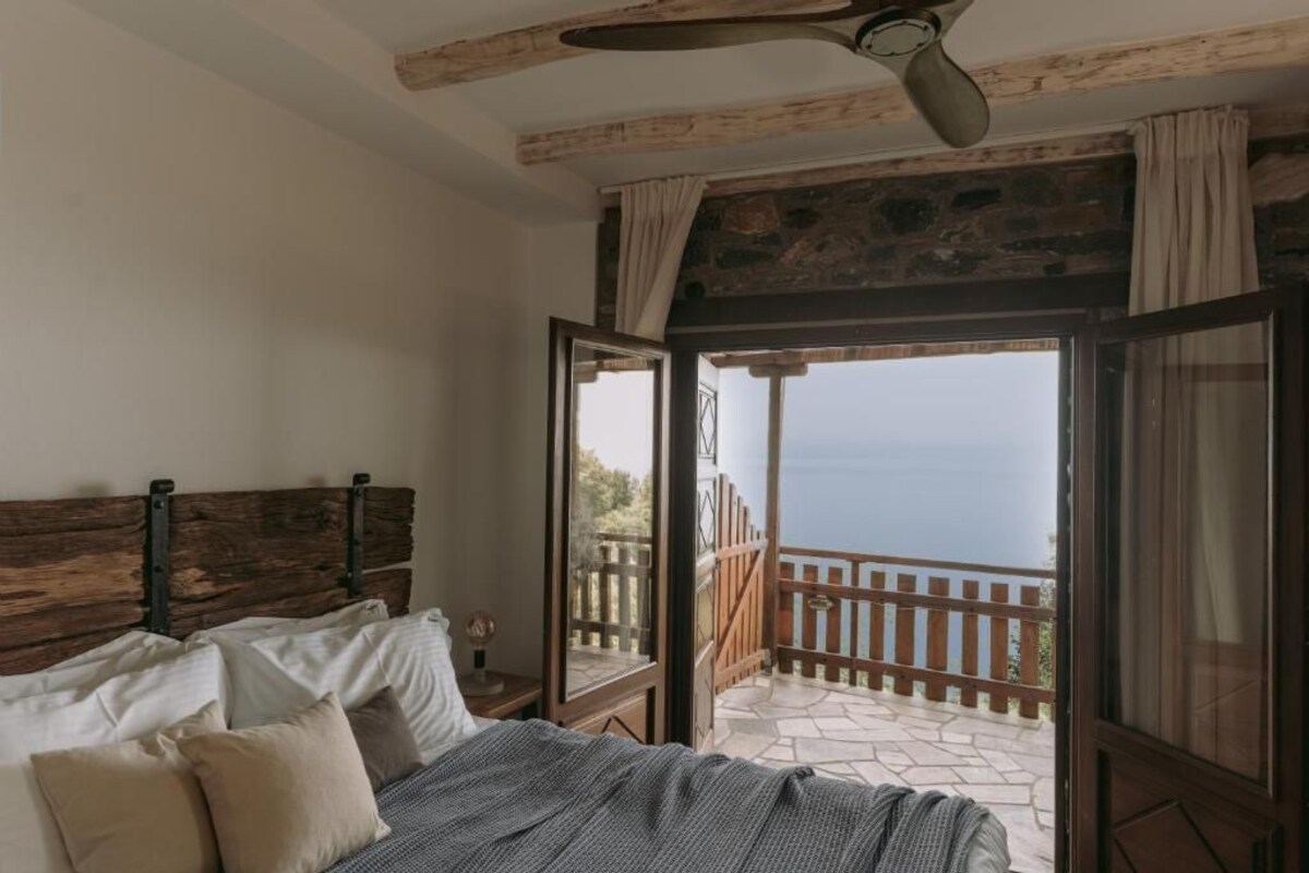 A bedroom with a wooden headboard, a bed with pillows, a window with curtains, and a view of the sea.