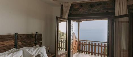 Un dormitorio con cabecera de madera, cama con almohadas, ventana con cortinas y vista al mar.