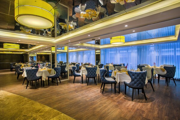 A spacious dining area with multiple tables and chairs, high ceilings with decorative elements, and large windows with curtains.