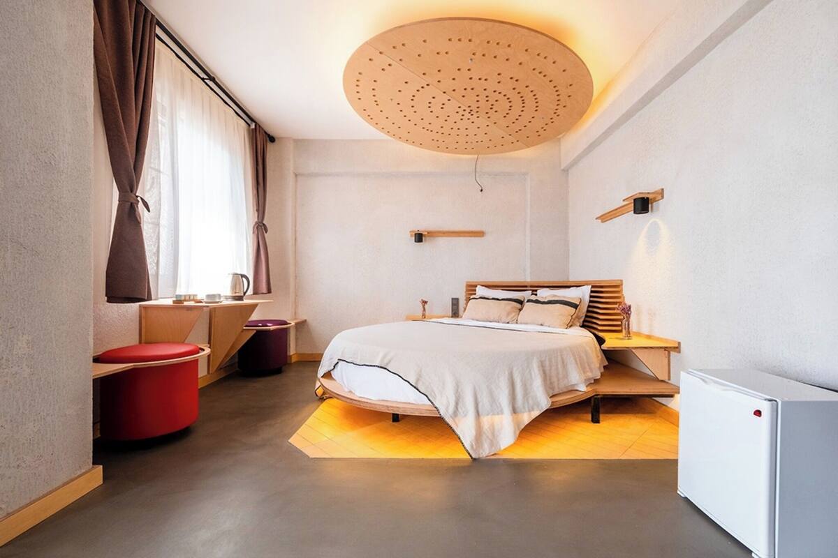 Ein modernes Hotelzimmer mit einem Bett, einem Schreibtisch, einem roten Sessel, einem kleinen Tisch und einem weißen Schrank.