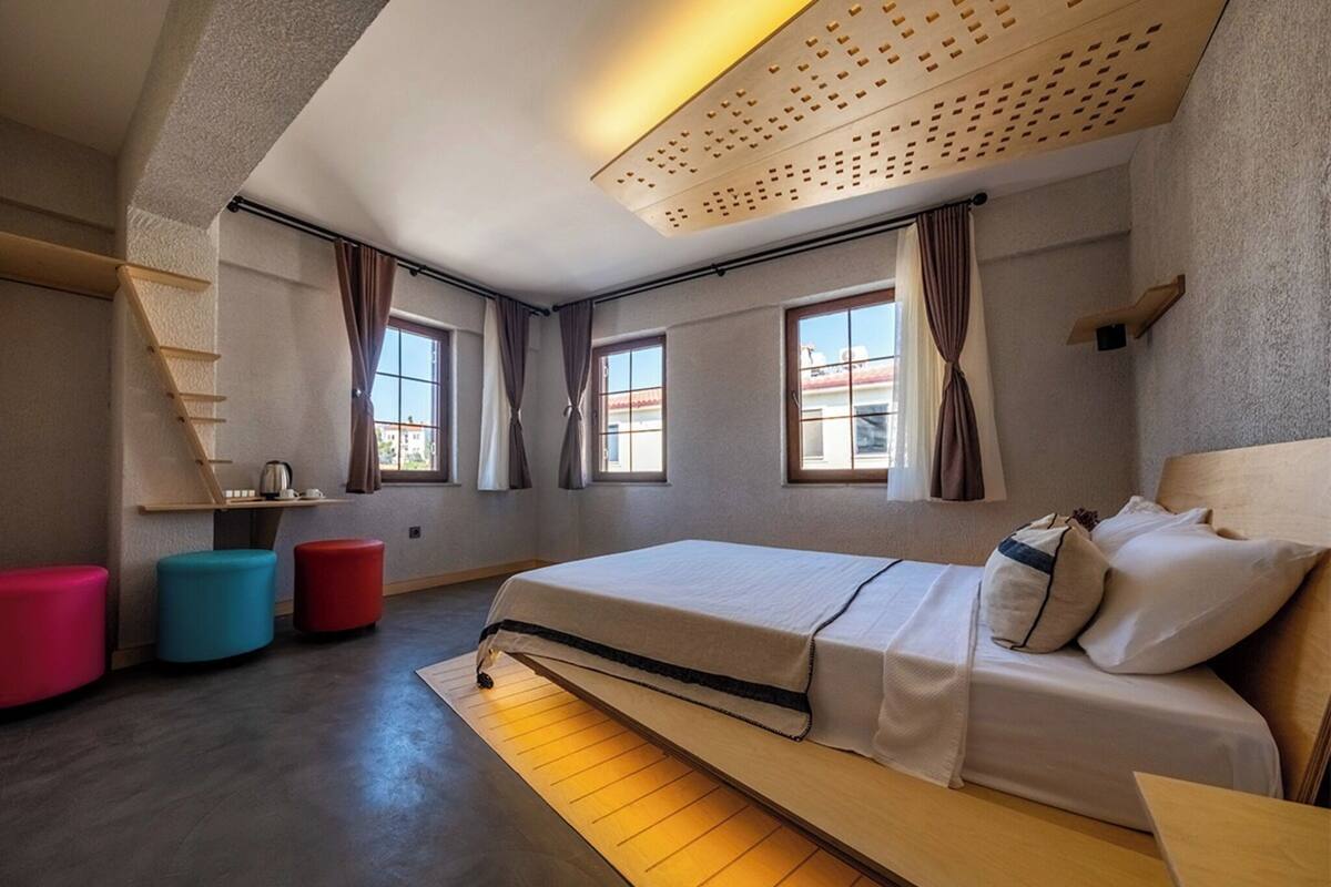 Ein modernes Schlafzimmer mit einem Bett, Fenstern, einer Holzleiter und bunten Hocker.