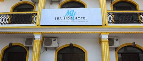 A hotel facade with a sign reading 'MY SEA SIDE HOTEL,' featuring a balcony with decorative railings and multiple air conditioning units.