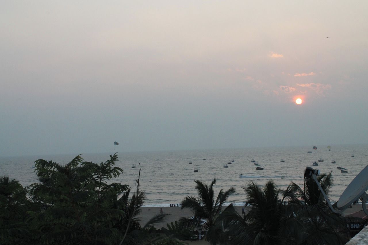 Vista da praia com palmeiras, um pôr do sol e barcos na água.