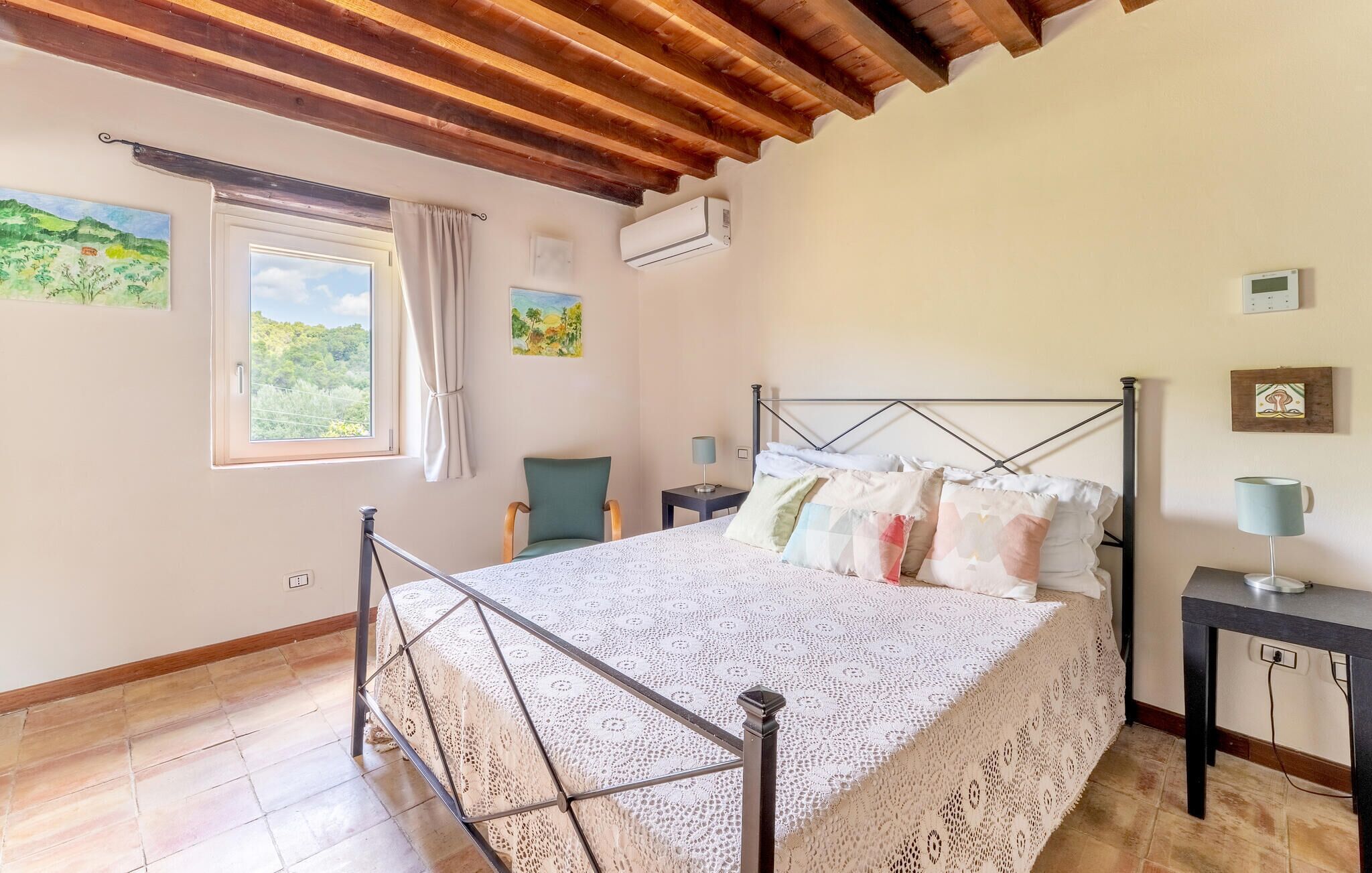 Un dormitorio con cama, ventana con cortinas, una silla, mesita de noche con lámpara y obras de arte en las paredes.