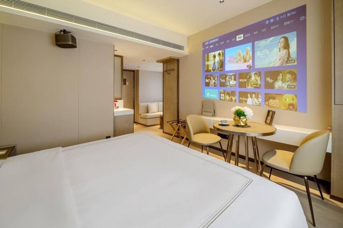 Habitación de hotel con una cama grande, un escritorio con sillas, una mesita con un florero y un televisor de pared que muestra carteles de películas.