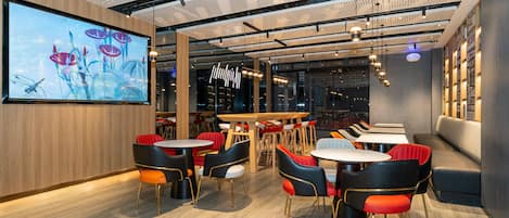A modern restaurant interior with wooden flooring, multiple dining tables and chairs, a large screen displaying an image, and a ceiling with a perforated design.