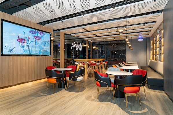 A modern restaurant interior with wooden flooring, multiple dining tables and chairs, a large screen displaying an image, and a ceiling with a perforated design. - Ibis Xian Bell Tower Huimin Street (Xi'an)