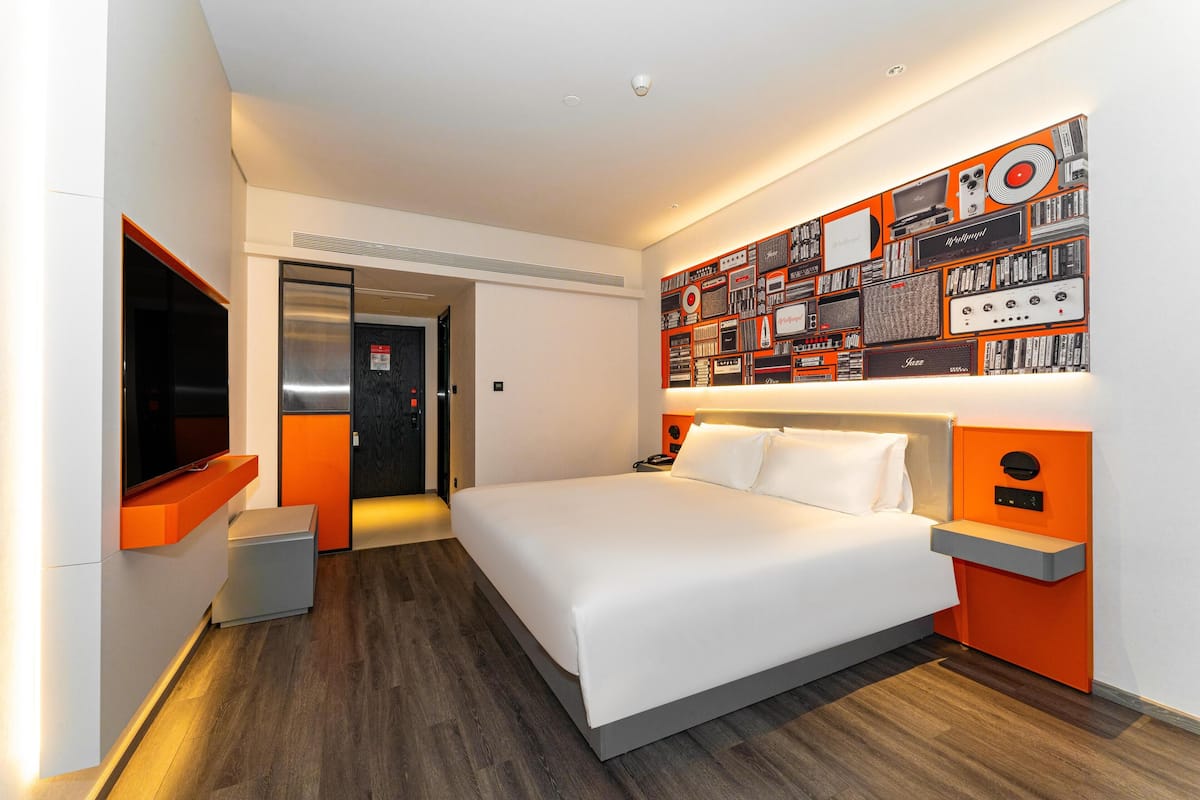 A modern hotel room with a large bed, a flat-screen TV, a wall-mounted sound system, and a door leading to the bathroom.