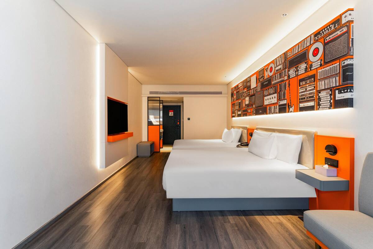 A modern hotel room with a large bed, a television, a wall-mounted shelf with books, and a wooden floor.