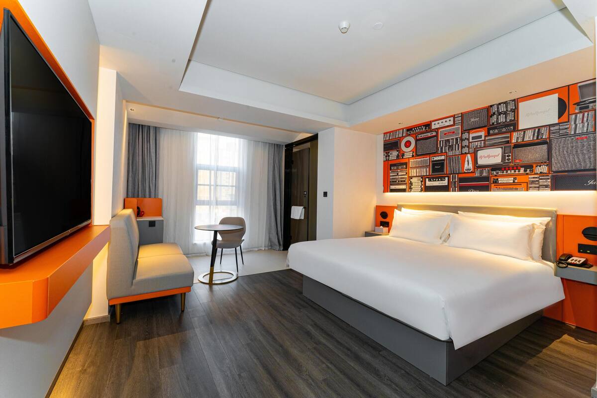 A hotel room with a large bed, a flat-screen TV, a seating area with a chair and table, and a wall decorated with vintage audio equipment.