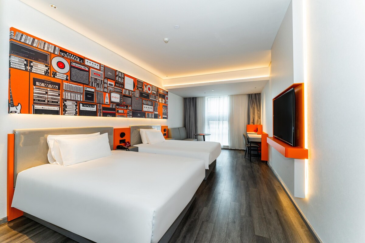 Habitació d'hotel amb dos llits, un televisor, una taula de treball i una paret amb diversos objectes.