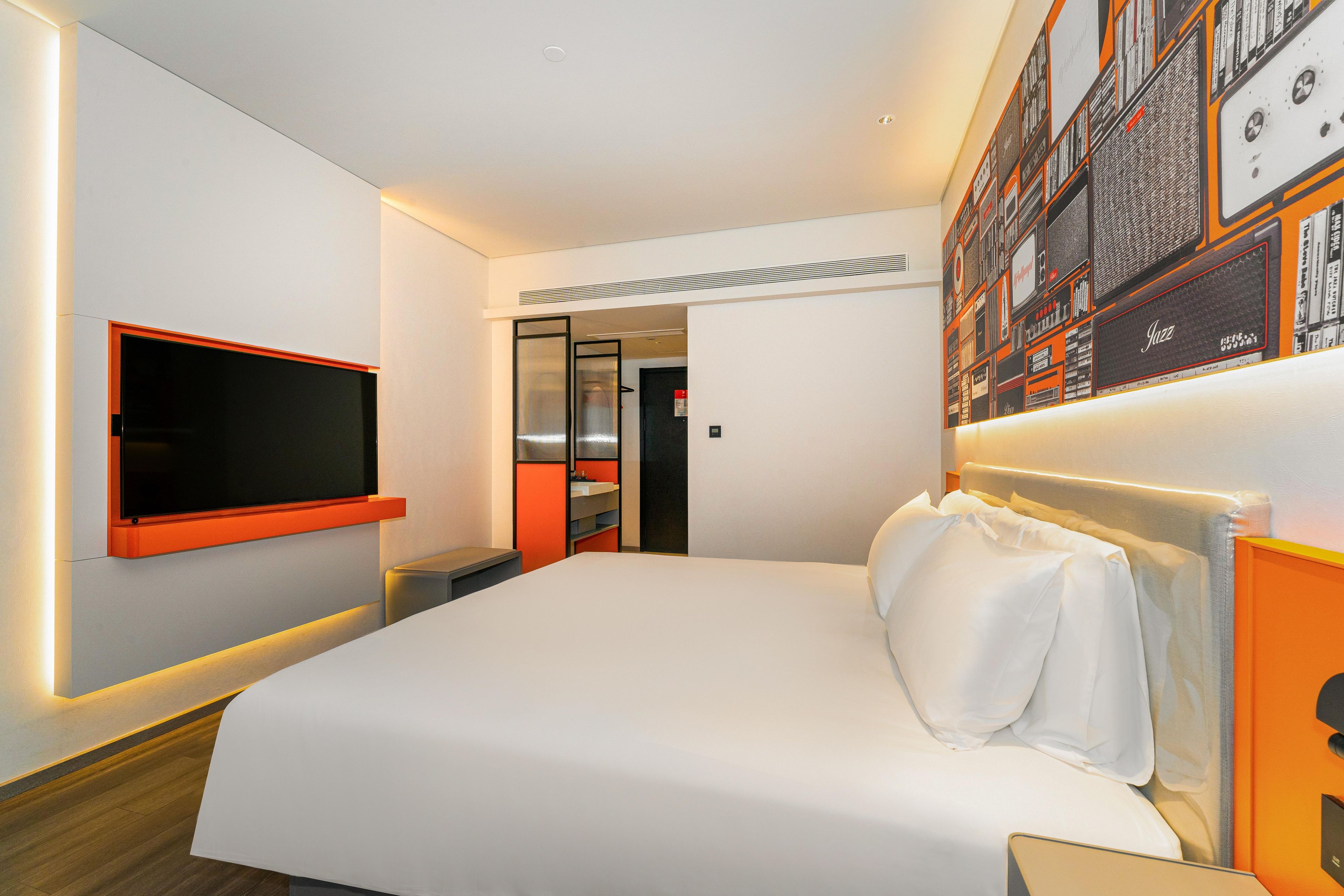 Una habitación de hotel moderna con una cama grande, un televisor de pantalla plana en la pared y una estantería con varios objetos.