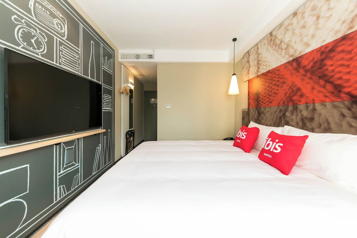 A hotel room with a large flat-screen TV, a bed with red pillows, and a wall with abstract designs.