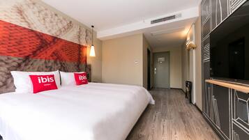 A hotel room with a large bed, a television, a wooden floor, and a hallway leading to a door.