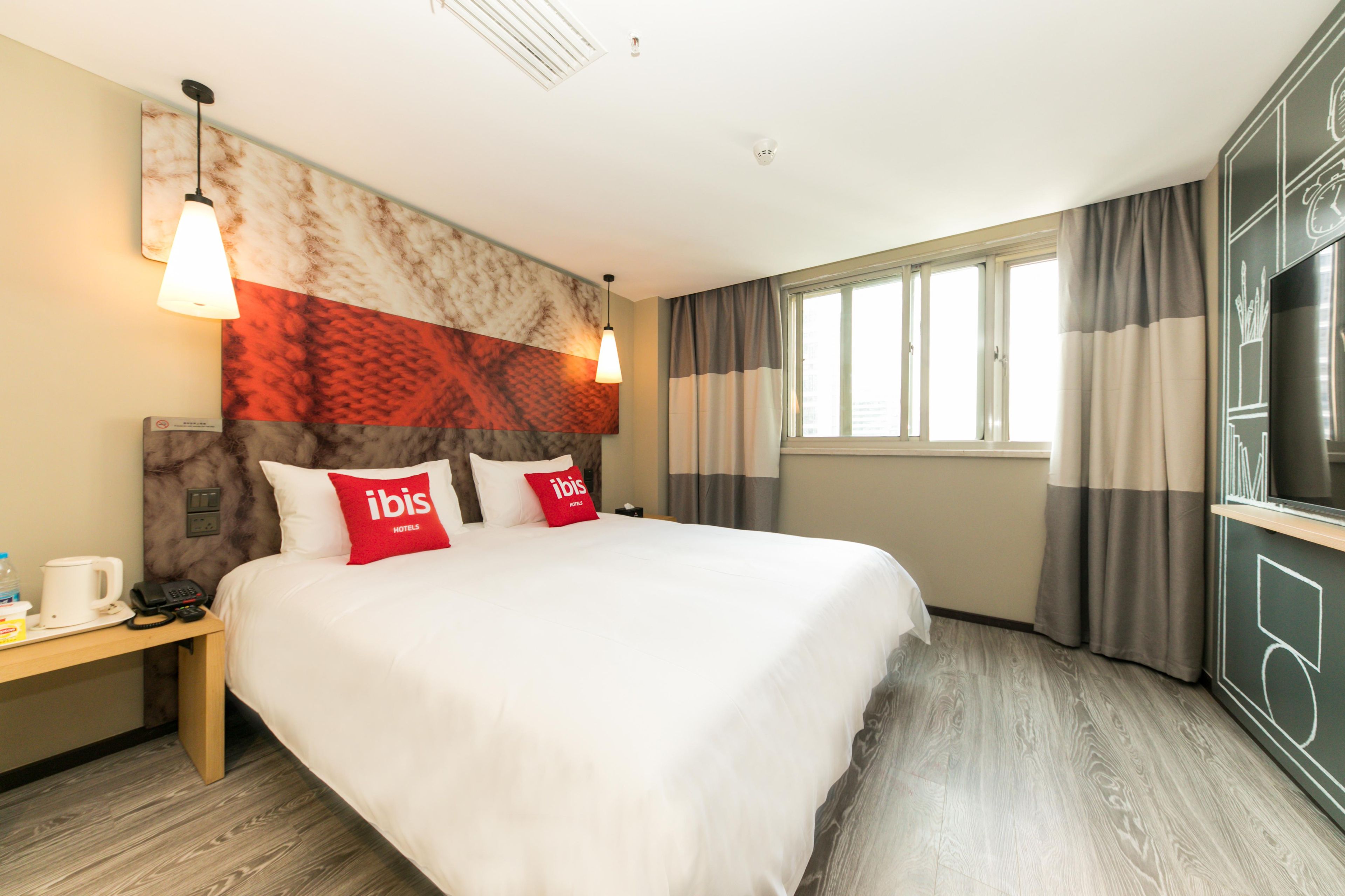 Une chambre d’hôtel avec un grand lit, deux oreillers rouges, une table de chevet avec un téléphone et une machine à café, une télévision et une fenêtre avec des rideaux.