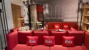 Un canapé rouge agrémenté de coussins à l’effigie de la marque « ibis HOTELS », un mur en bois et une cloison en verre.