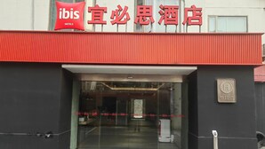 L’entrée d’un hôtel, avec un toit rouge et des portes en verre, une enseigne indiquant « ibis Hotels » et un logo en forme de « H » signalant la présence d’un hôtel.
