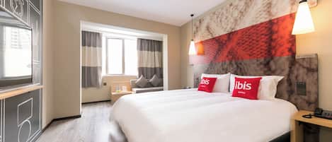 A hotel room with a large bed, a desk, a chair, a window with curtains, and a wall with a clock and abstract design.