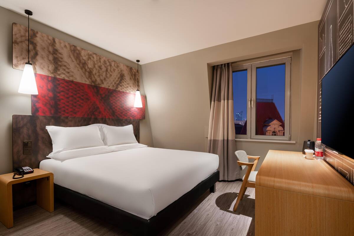 Ein Hotelzimmer mit Bett, Nachttisch mit Telefon, Schreibtisch mit Stuhl und einem großen Fenster mit Vorhängen.