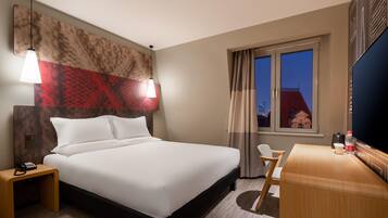 A hotel room with a bed, bedside table with a phone, a desk with a chair, and a large window with curtains.