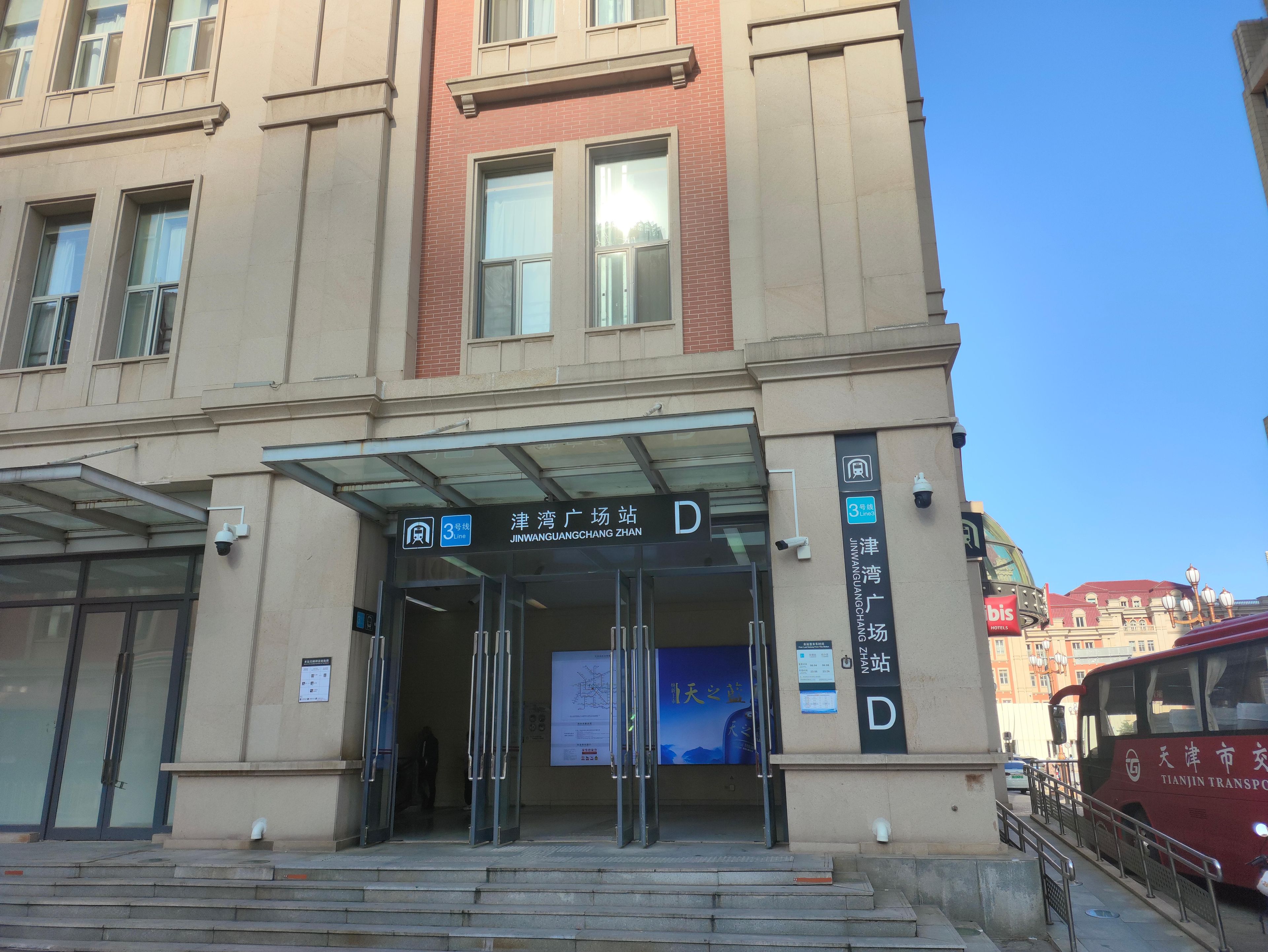Entrada de um edifício com toldo de vidro, sinalização com 'JINWANGUANGZHANG ZHAN' e placa de ponto de ônibus.