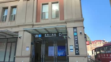 Entrada de um edifício com toldo de vidro, sinalização com 'JINWANGUANGZHANG ZHAN' e placa de ponto de ônibus.