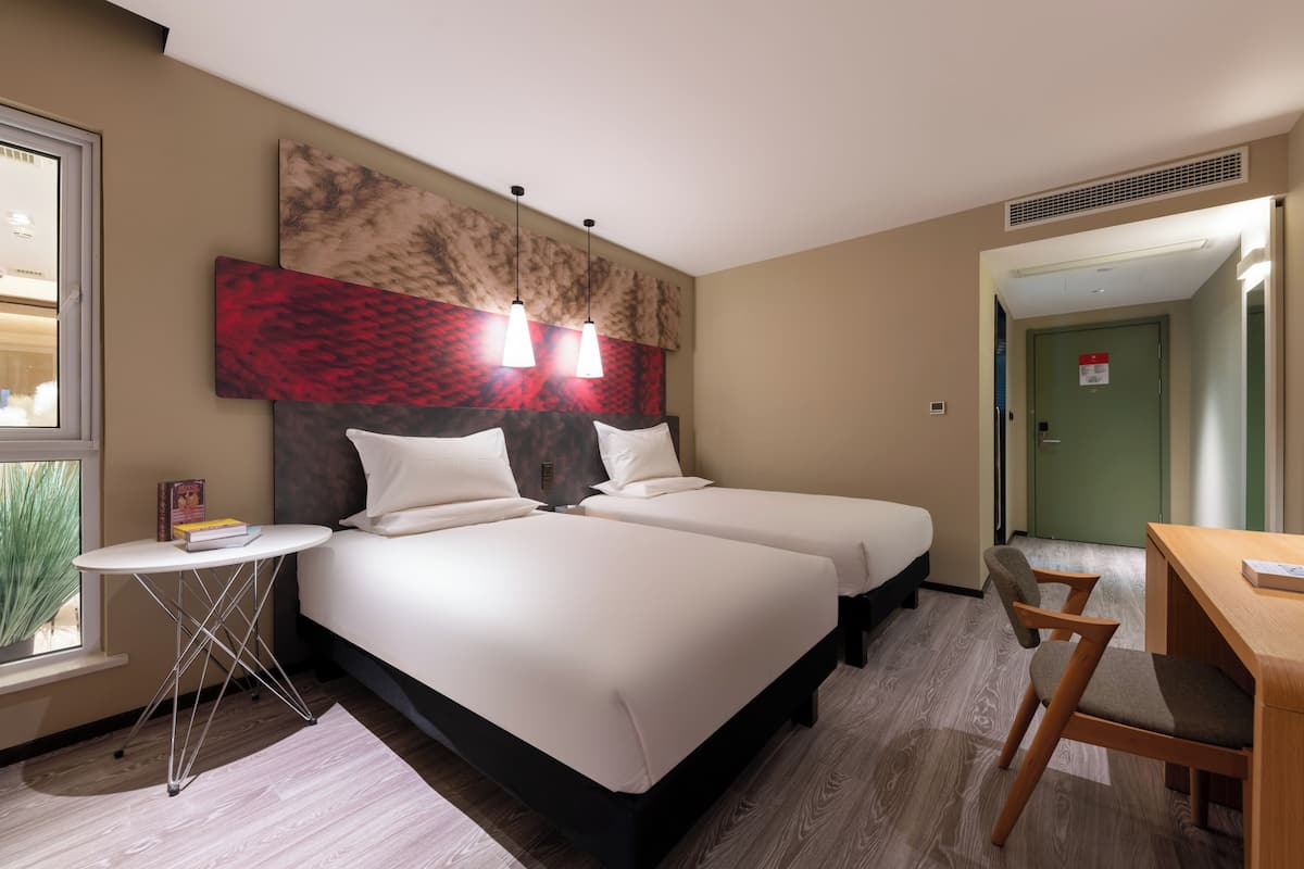 Hotellihuoneessa on kaksi sänkyä, pieni pöytä, tuoli ja suuri, abstrakti taulu seinällä.