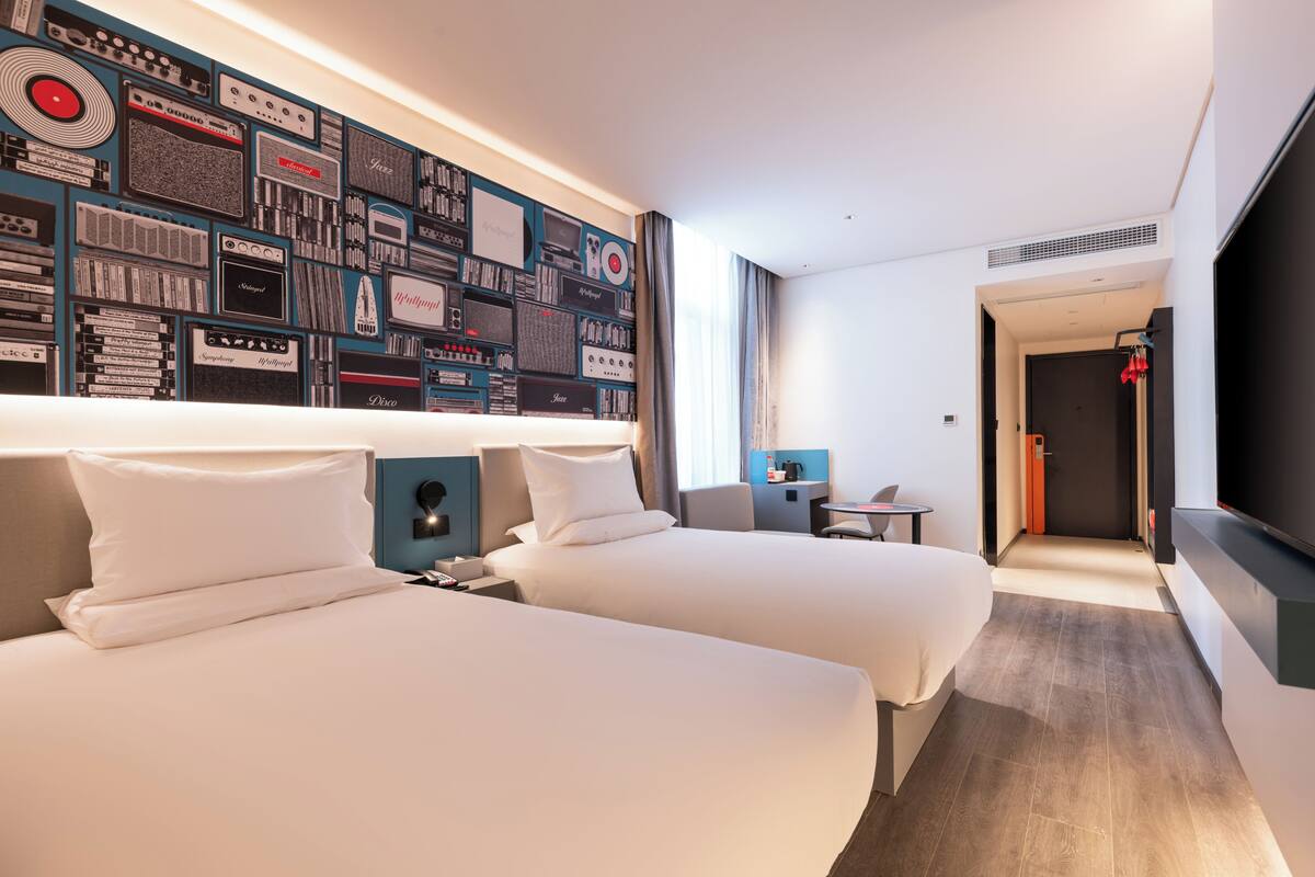 Ein Hotelzimmer mit zwei Betten, einer Wand voller Vintage-Audioequipment, einem Fernseher und einem kleinen Schreibtisch.