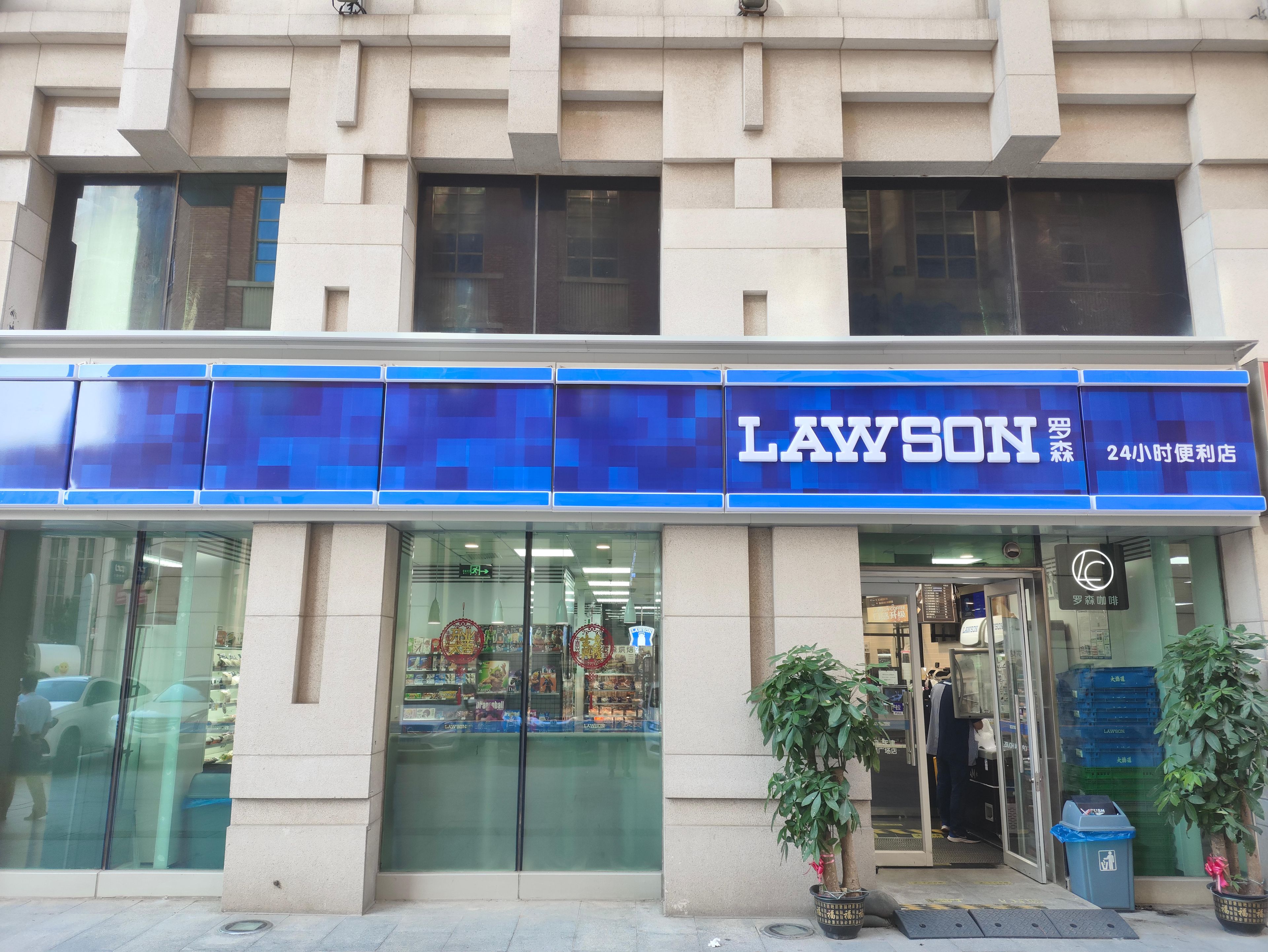 Un magasin LAWSON avec une enseigne bleue, une vitrine en verre et une plante en pot à l’extérieur.