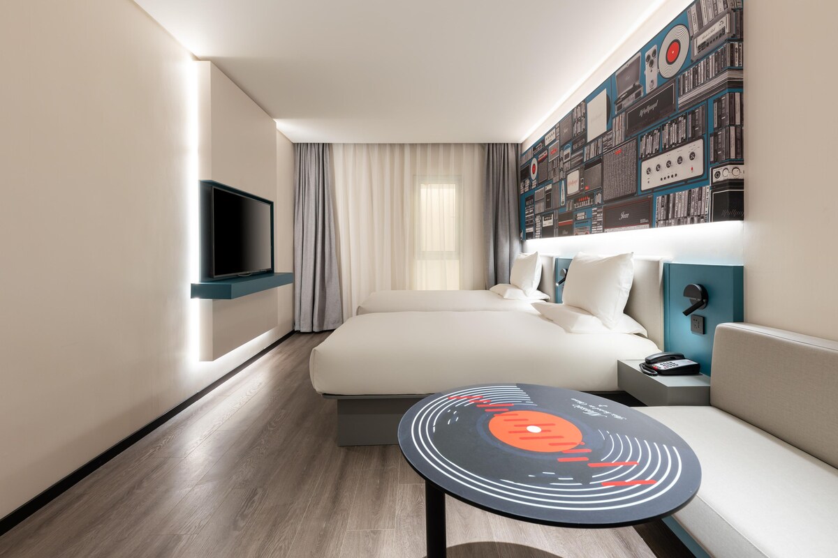 Ein modernes Hotelzimmer mit einem Bett, einem Flachbildfernseher, einem kleinen Tisch mit Vinyl-Schallplatten-Design und einer Wand, die mit Vintage-Audioausrüstung dekoriert ist.