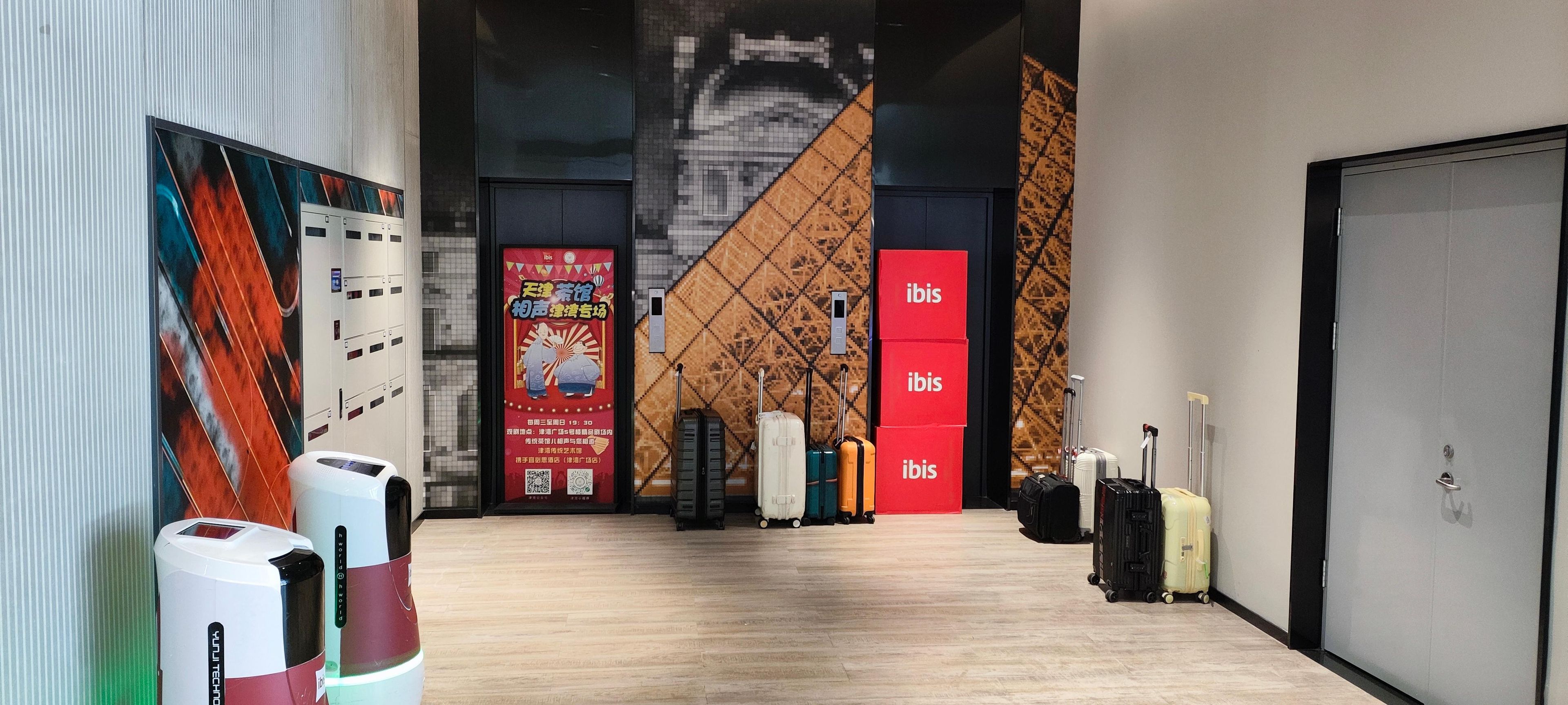 Un vestíbulo de hotel con diseño moderno, que cuenta con una gran pantalla digital, un portaequipajes y un llamativo letrero 'ibis'.