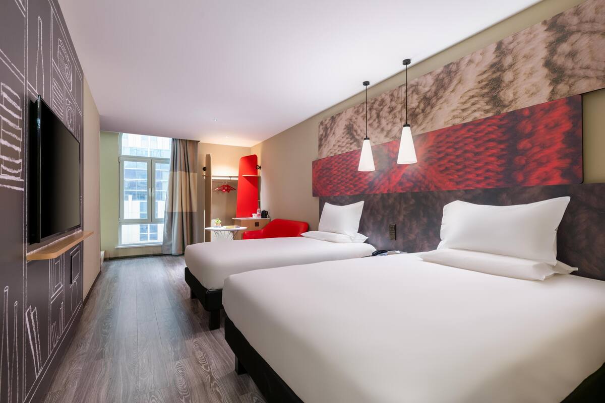 Ein Hotelzimmer mit zwei Betten, einem Fernseher, einem roten Sessel, einem Schreibtisch und einem großen Fenster.