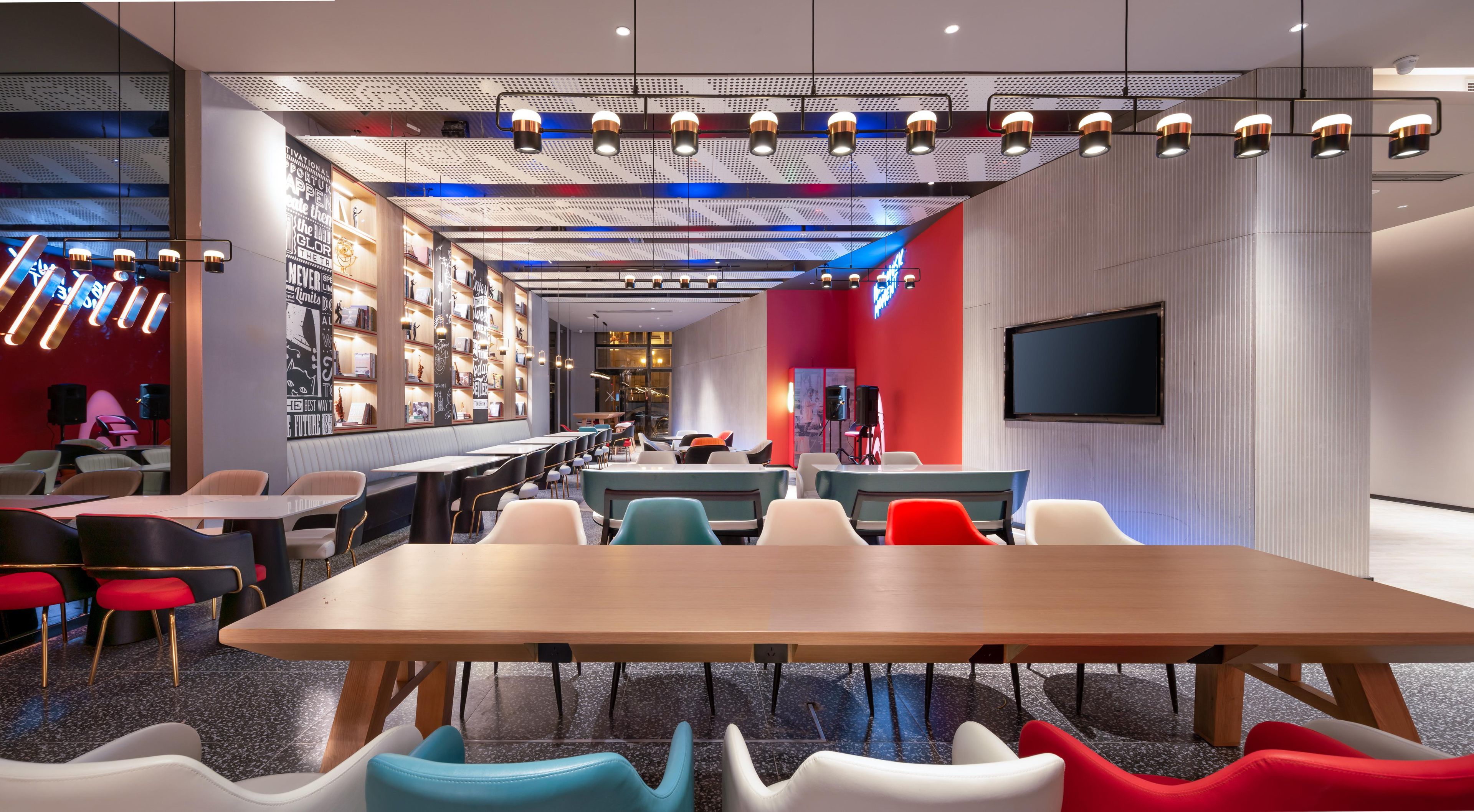 Un restaurante moderno con una larga mesa de madera, sillas coloridas y una zona de bar con estanterías.