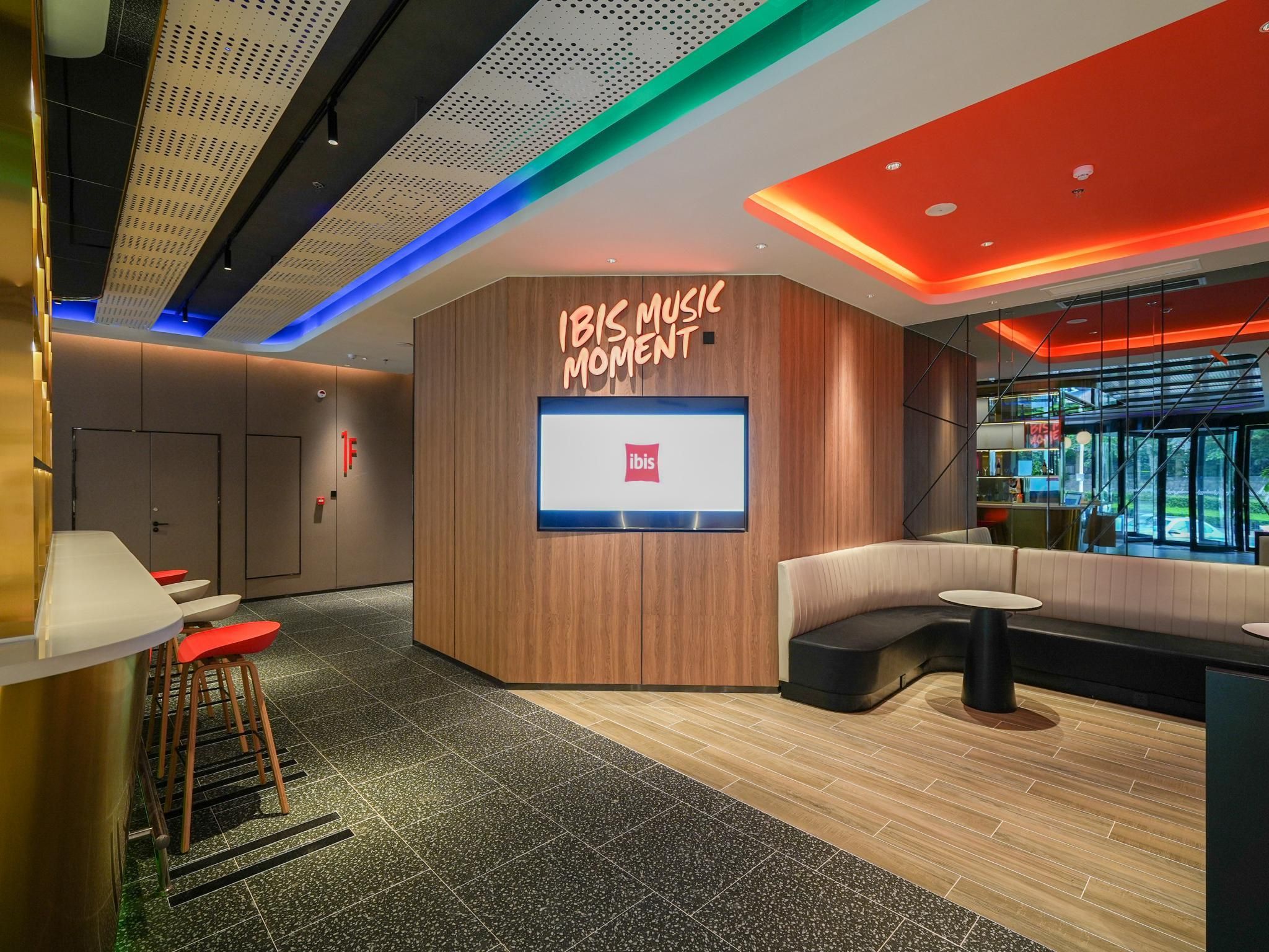 Ein modernes Interieur mit einer Sitzecke, einem digitalen Bildschirm mit dem ibis-Logo und einer Wand als Holzdekor.