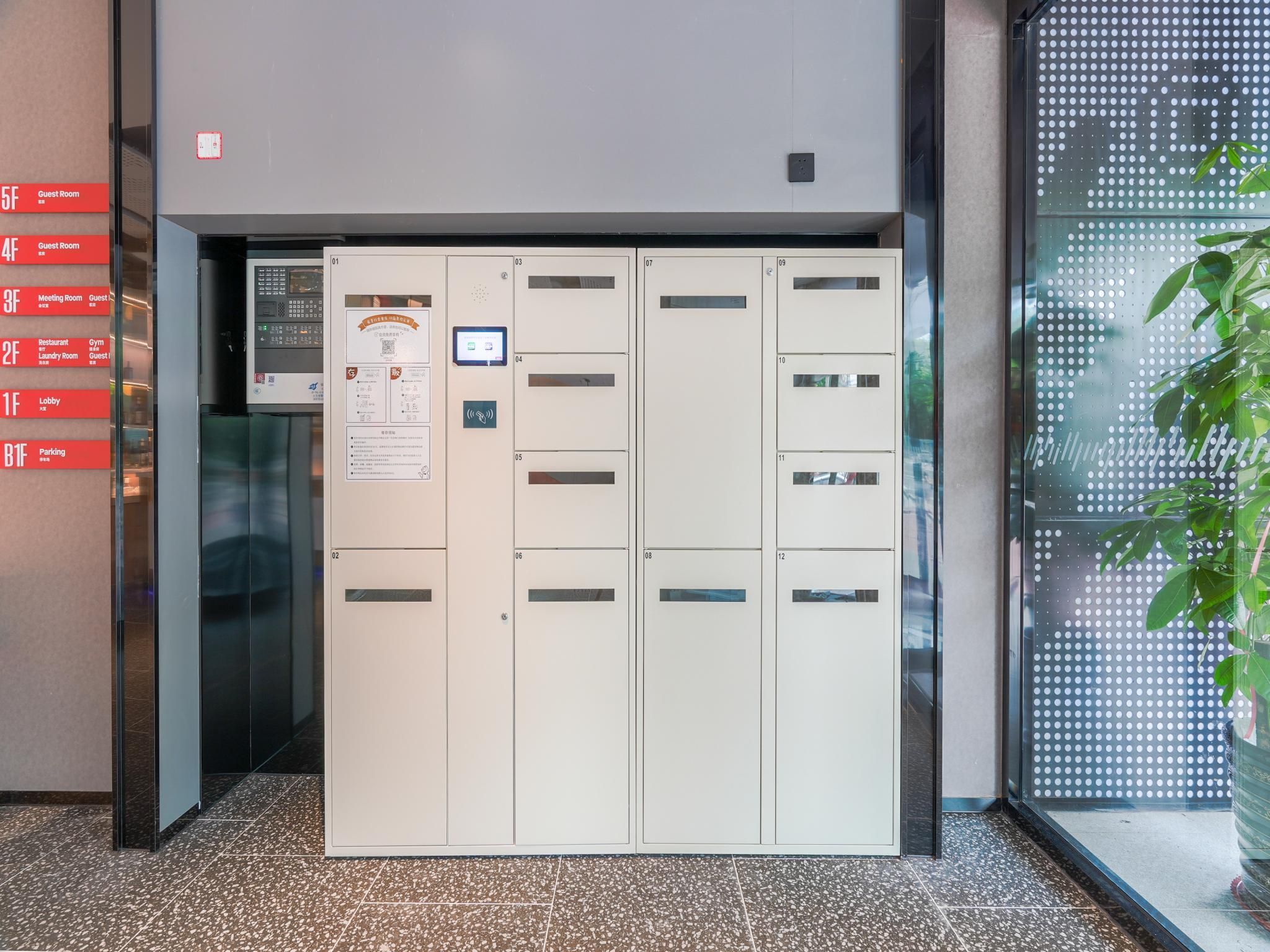 A hotel entrance with a set of lockers, a digital information board, and a glass door with a view of the outside.