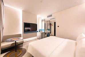 A hotel room with a bed, a TV, a small table, a sofa, and a bathroom with a mirror and sink. - Ibis Qingdao Xinduxin Capitamall Hotel (Qingdao)