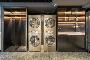 A modern laundry facility with two wall-mounted washing machines, a stainless steel sink, and a security camera. - Ibis Qingdao Xinduxin Capitamall Hotel (Qingdao)