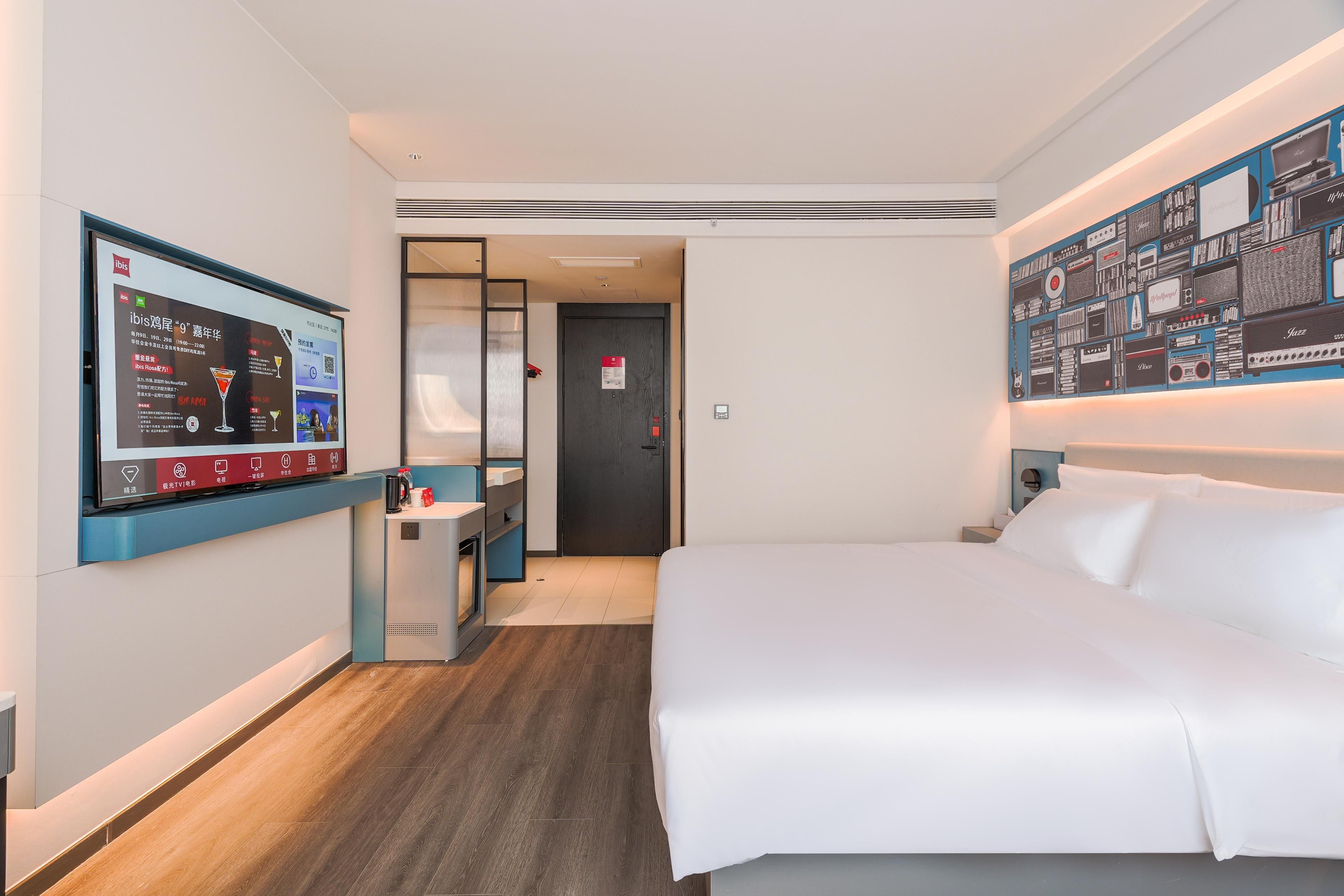 A hotel room with a bed, a TV mounted on the wall, a desk with a chair, and a door leading to another room.