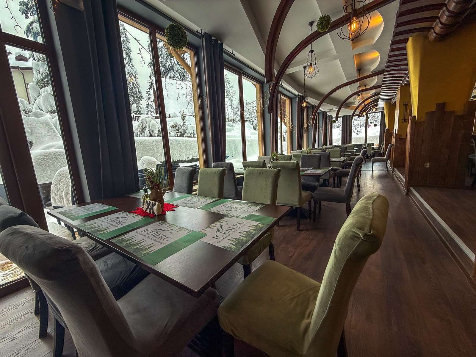 A restaurant with tables and chairs, large windows with views of snow-covered trees, and a wooden ceiling.