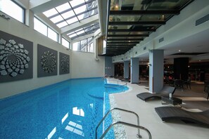 An indoor swimming pool with a glass ceiling, lounge chairs, and decorative wall art. - Hotel Iceberg (Sofia)