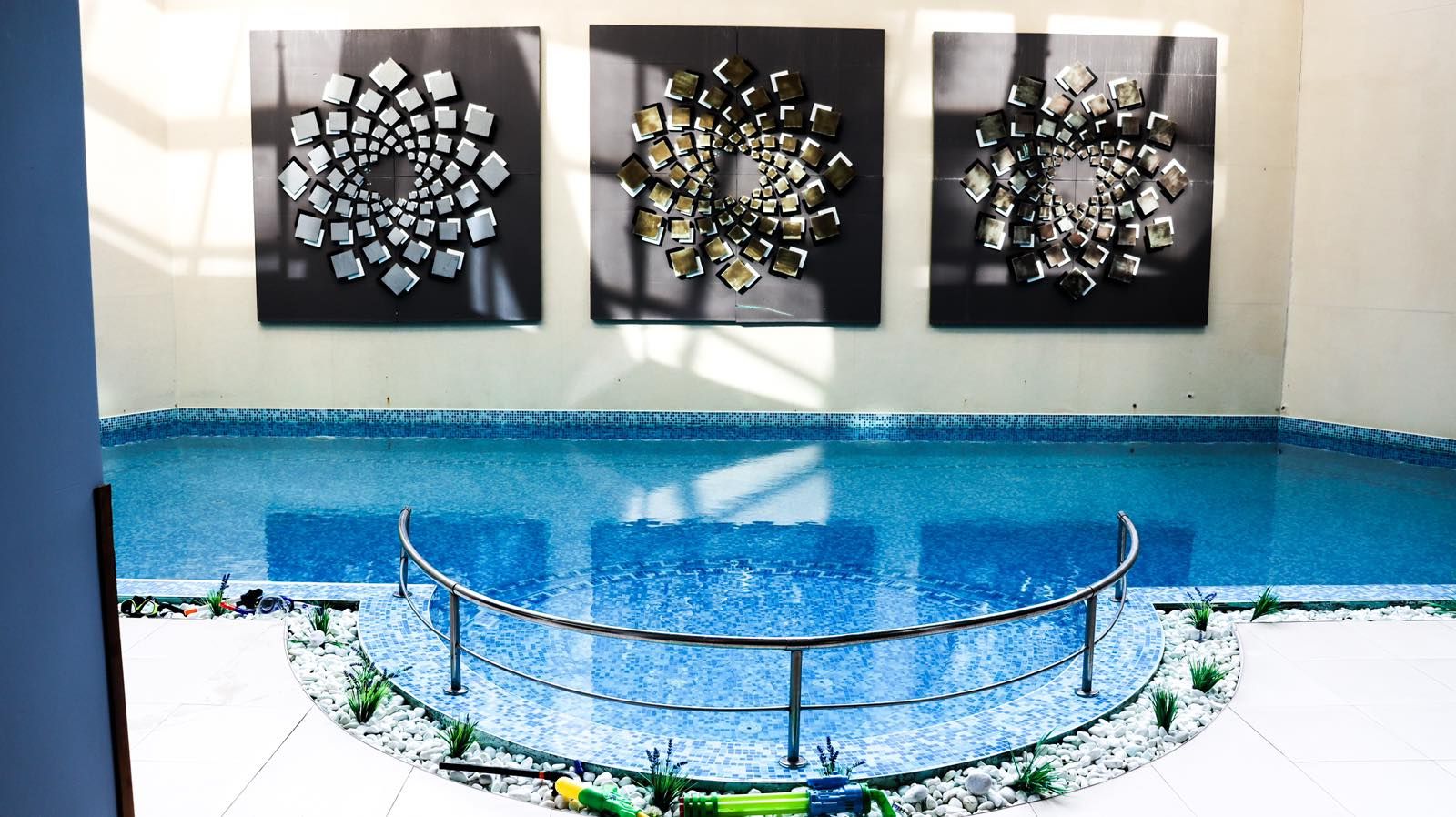 Una piscina interior con borde de azulejos, rodeada por una barandilla metálica y tres piezas de arte abstracto en la pared.