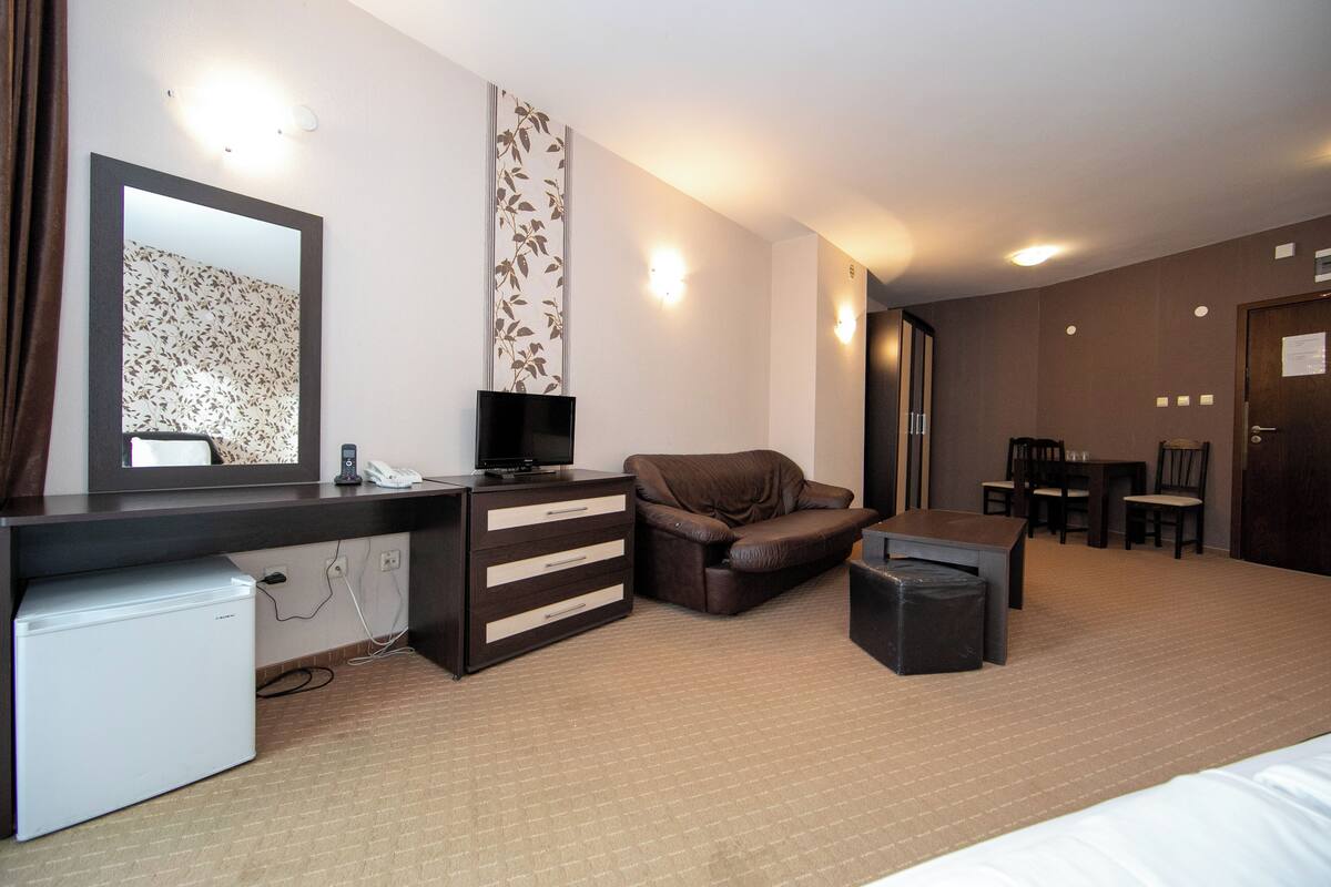A hotel room with a bed, a desk with a computer, a chair, a small table, a sofa, a trash bin, and a television.