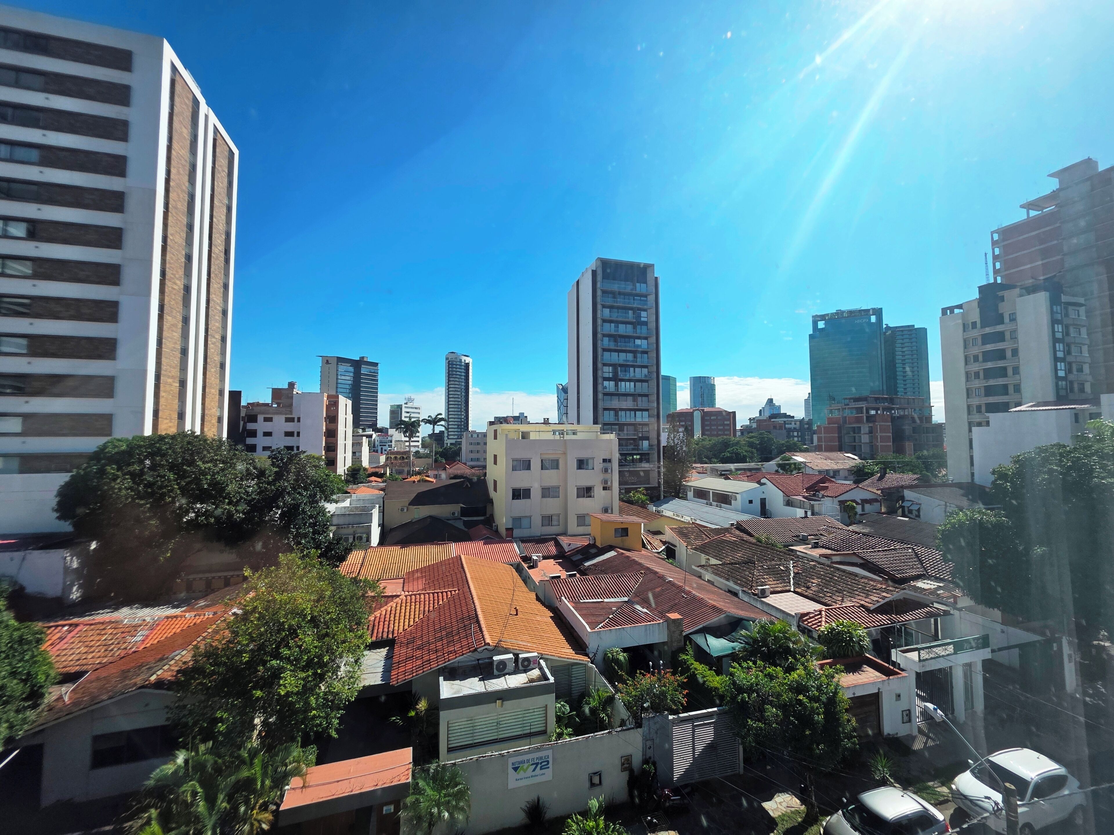Un paysage urbain mêlant des immeubles modernes de grande hauteur et des bâtiments traditionnels plus petits, un ciel bleu dégagé et un soleil éclatant.