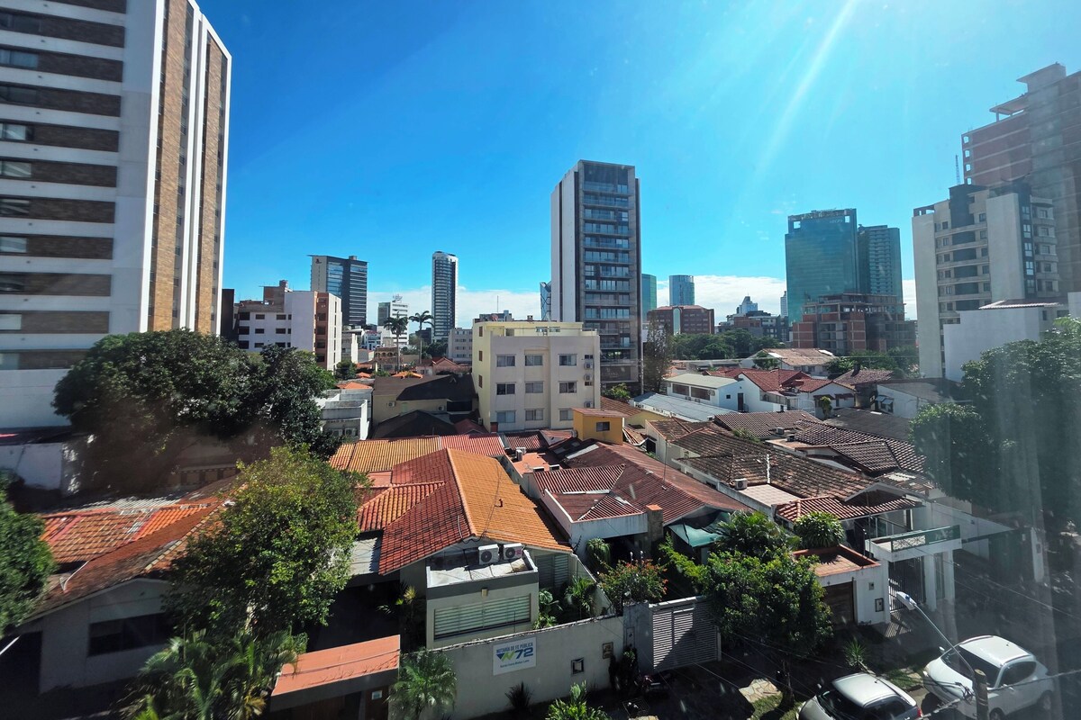 A cityscape with a mix of modern high-rise buildings and traditional low-rise structures, a clear blue sky, and a bright sun.